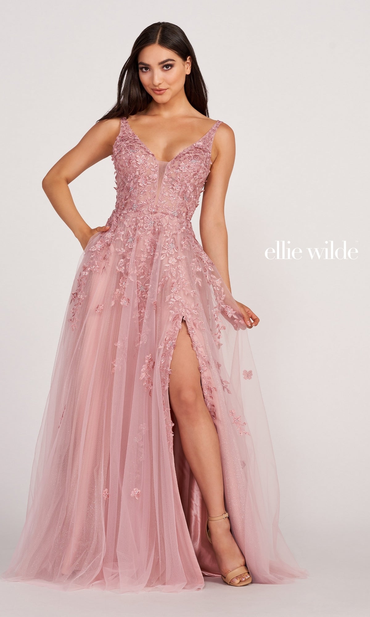 A woman wears the Ellie Wilde Side-Slit Long Prom Ball Gown EW34103 in blush pink with lace, a plunging neckline, floral appliqués, sleeveless design, and a thigh-high slit. The embellished dress is shown against a plain white background.